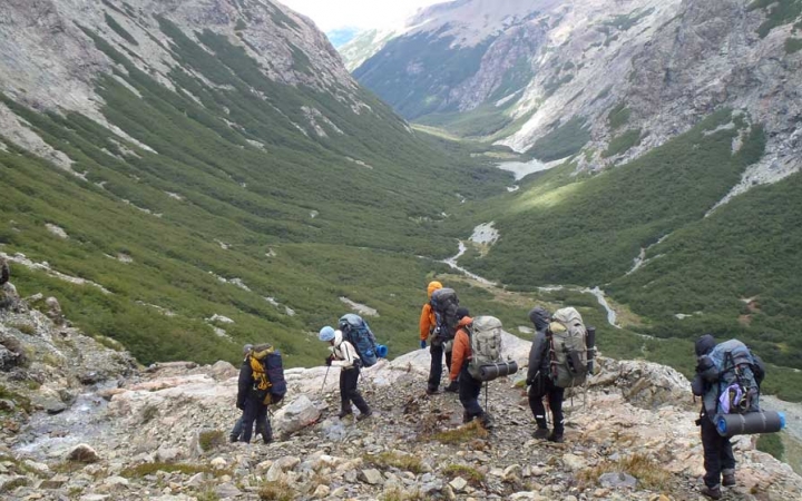 A group of people wearing backpacks hike over a rocky cliff, overlooking a vast green valley surrounded by mountains. There is a narrow river winding through the valley. 